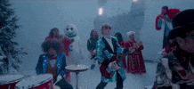 Music video gif. Ed Sheeran plays a guitar, strolling next to Elton John, playing the piano. Surrounded by snow and Christmas decor, musicians playing behind the artists in the music video, "Merry Christmas."