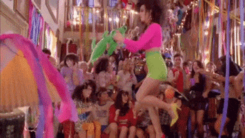 Music Video gif. Katy Perry is dancing on a table during her song, Last Friday Night. She shuffles as she moves her hands back and forth and the party around her goes wild.