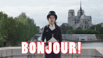 Video gif. A woman in a Victorian suit, complete with a top hat, stands on a bridge with a French Chateau in the background. She spreads her arms out to present the scenery and says, “Bonjour!”