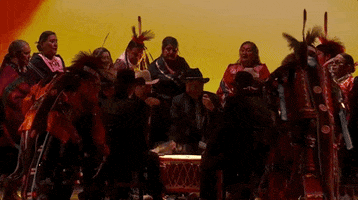 Video gif. A group of Native American dancers dressed in their traditional regalia dance in a circle around a large drum. They wear shades of red and black and shake their heads to the beat of the drum.