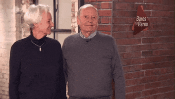 Video gif. An elderly man and woman stand side by side in front of a brick wall, smiling and nodding with wide eyes. Text, "Schauen wir mal was wird."